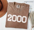 Folded brown (espresso) t-shirt with '2000' print on a white surface, accompanied by a hat and dried leaves.
