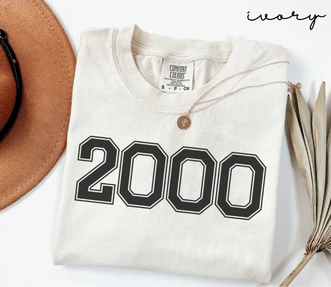 A beige (ivory) t-shirt with '2000' print, placed next to a brown hat and a dried plant on a light background.
