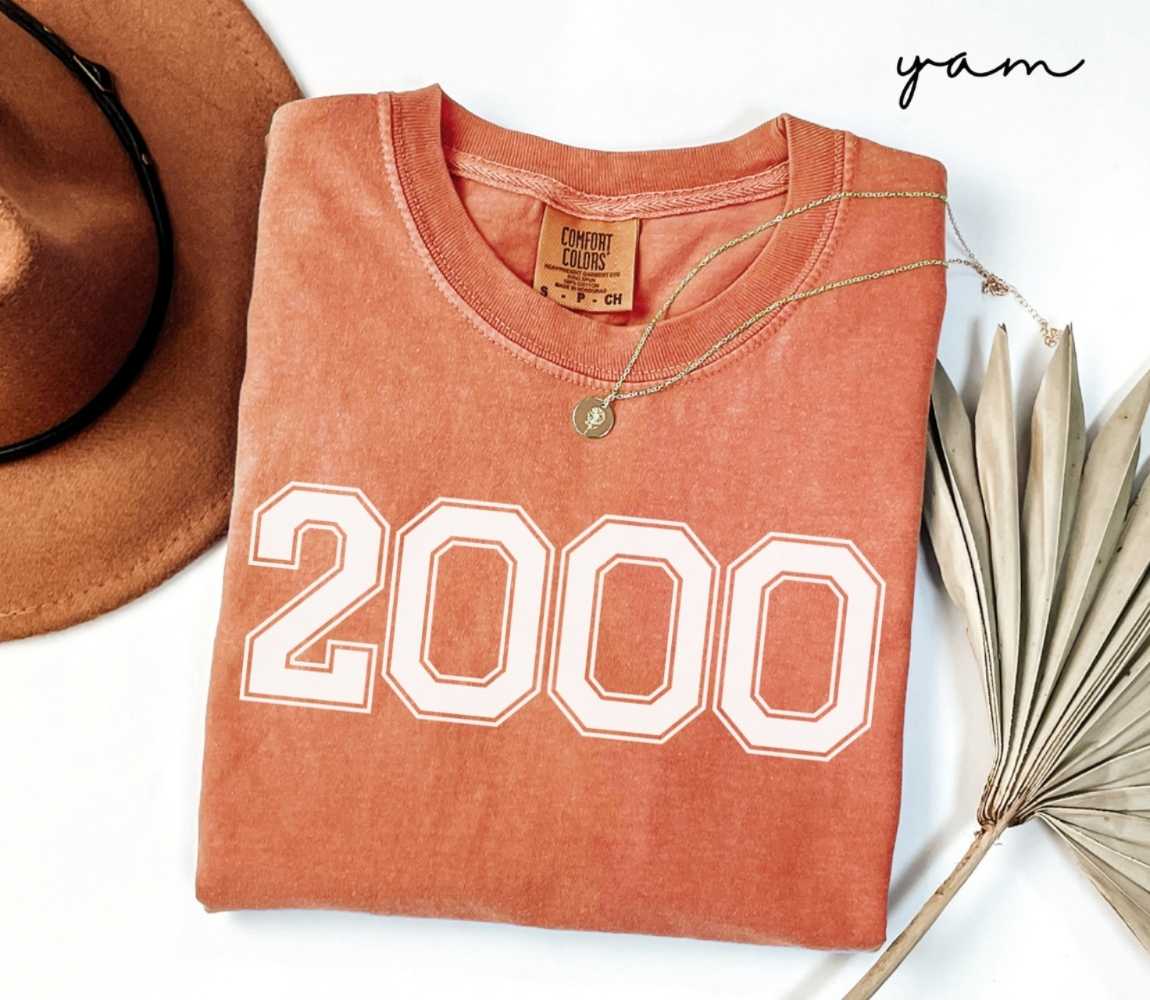 Folded orange (yam) t-shirt with a '2000' print, a brown hat, and a dried plant on a white background.
