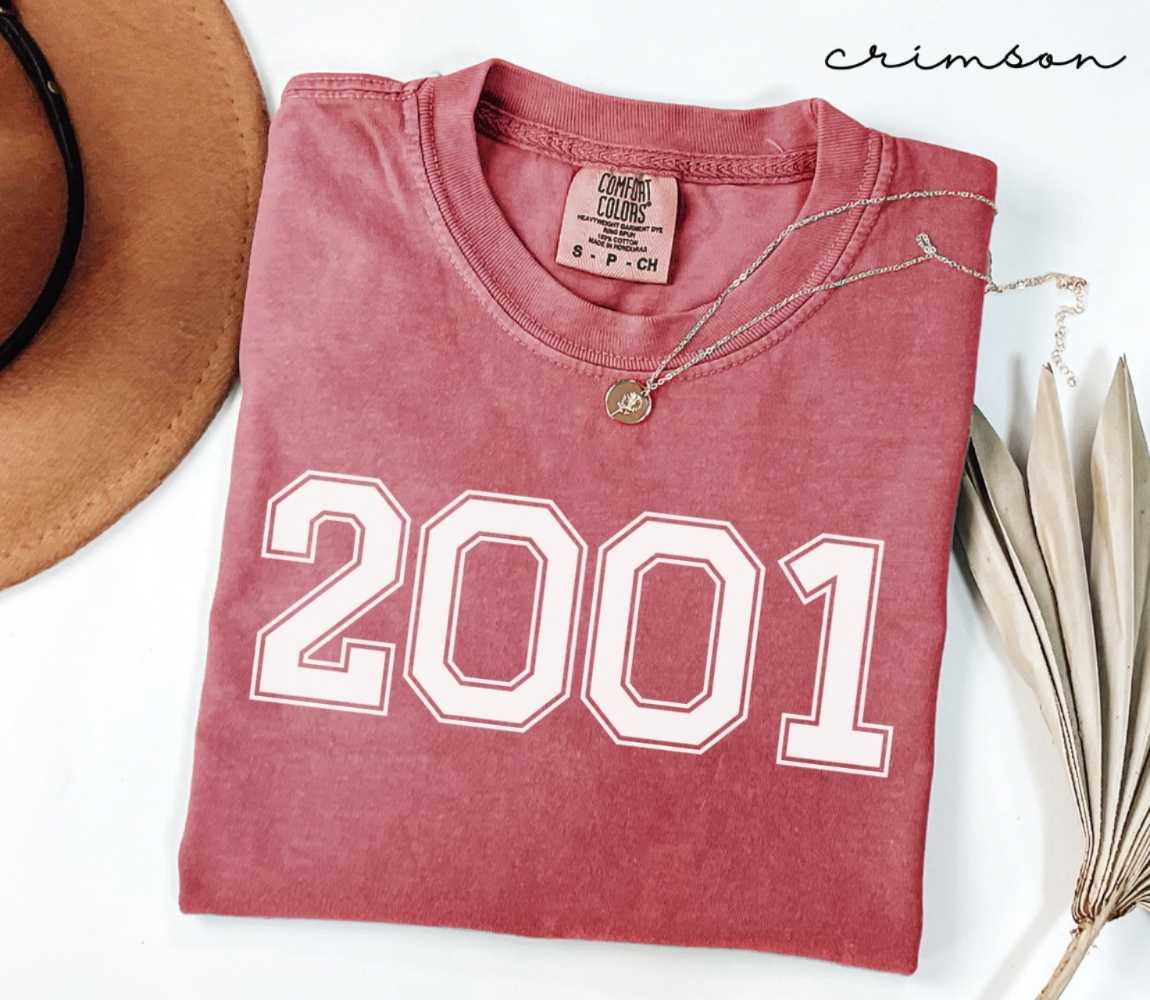 A red (Crimson) folded t-shirt with '2001' printed on it, next to a hat, and dried leaves on a white background.
