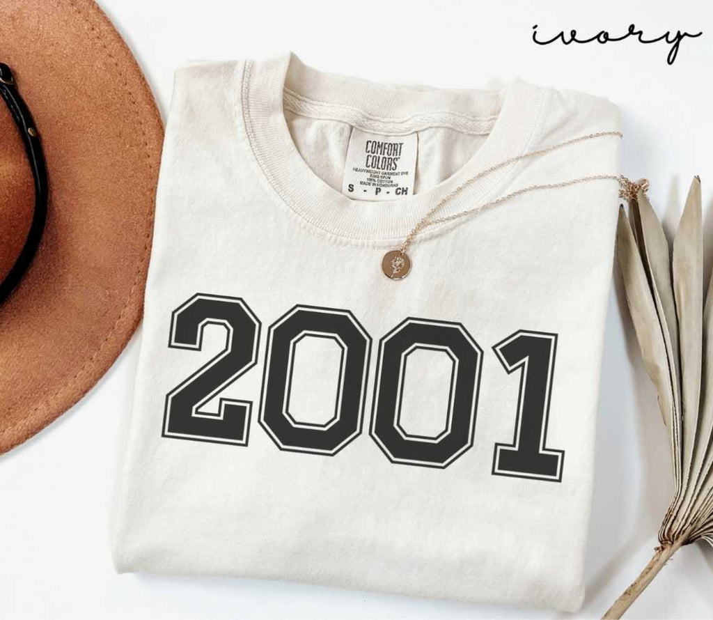 A beige (ivory) t-shirt with '2001' print, placed next to a brown hat and a dried plant on a light background.
