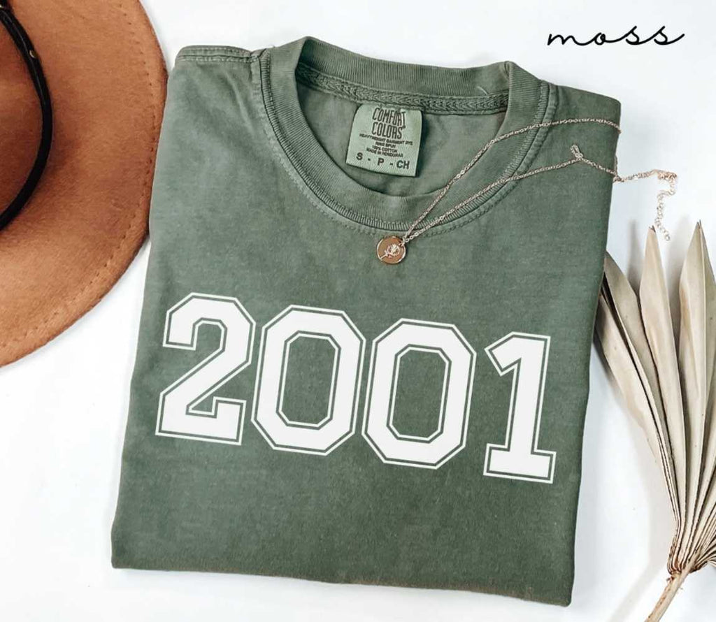 Folded green (moss) t-shirt featuring a '2001' print on a white surface alongside a hat and dried leaves.
