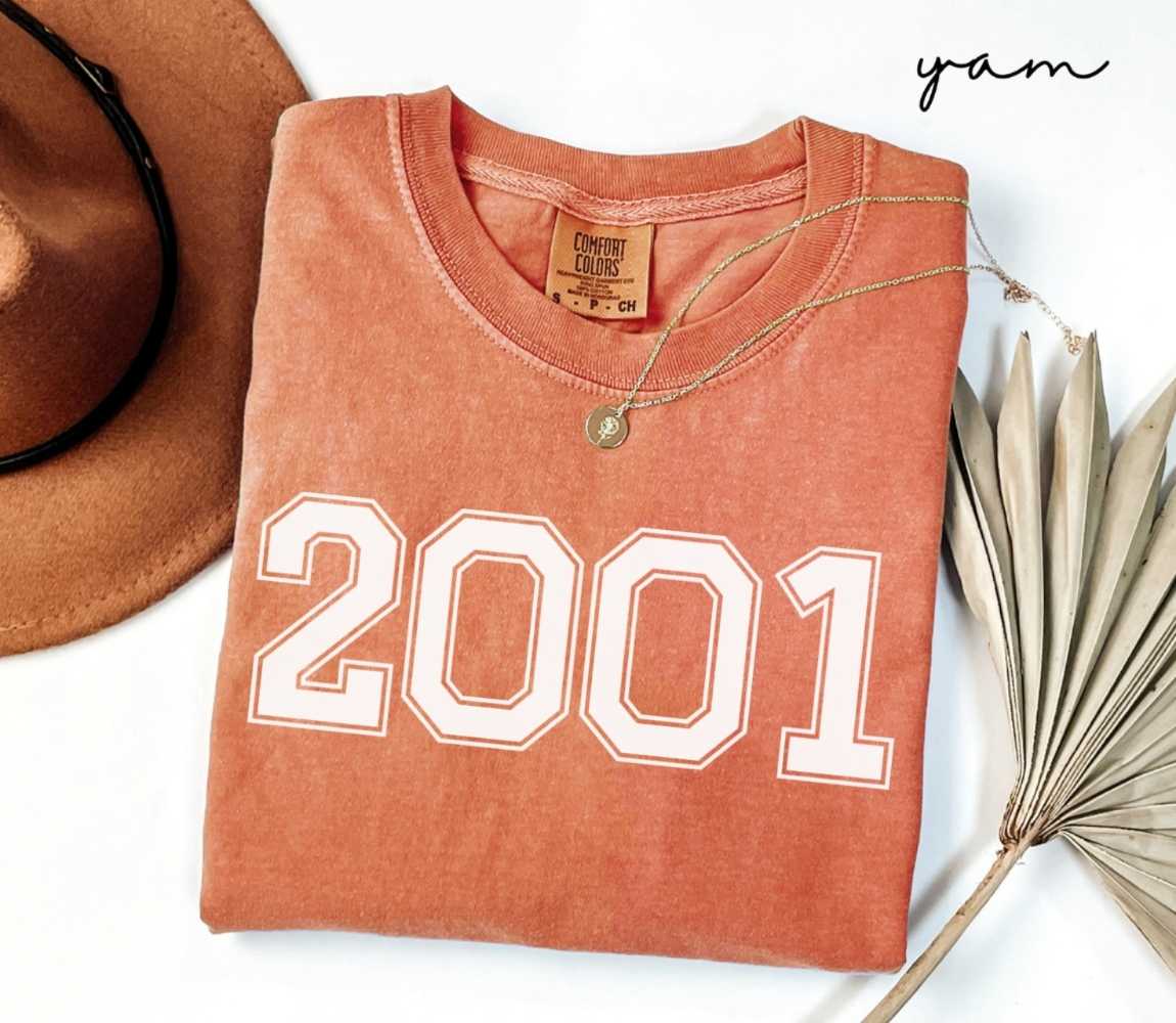 Folded orange (yam) t-shirt with a '2001' print, a brown hat, and a dried plant on a white background.
