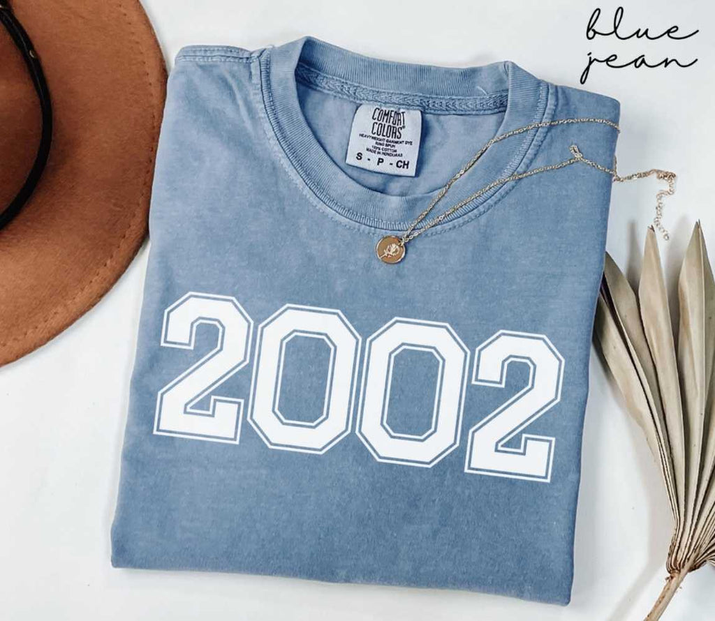 Folded blue jean t-shirt featuring '2002' print on a white surface, accompanied by a hat and dried leaves.
