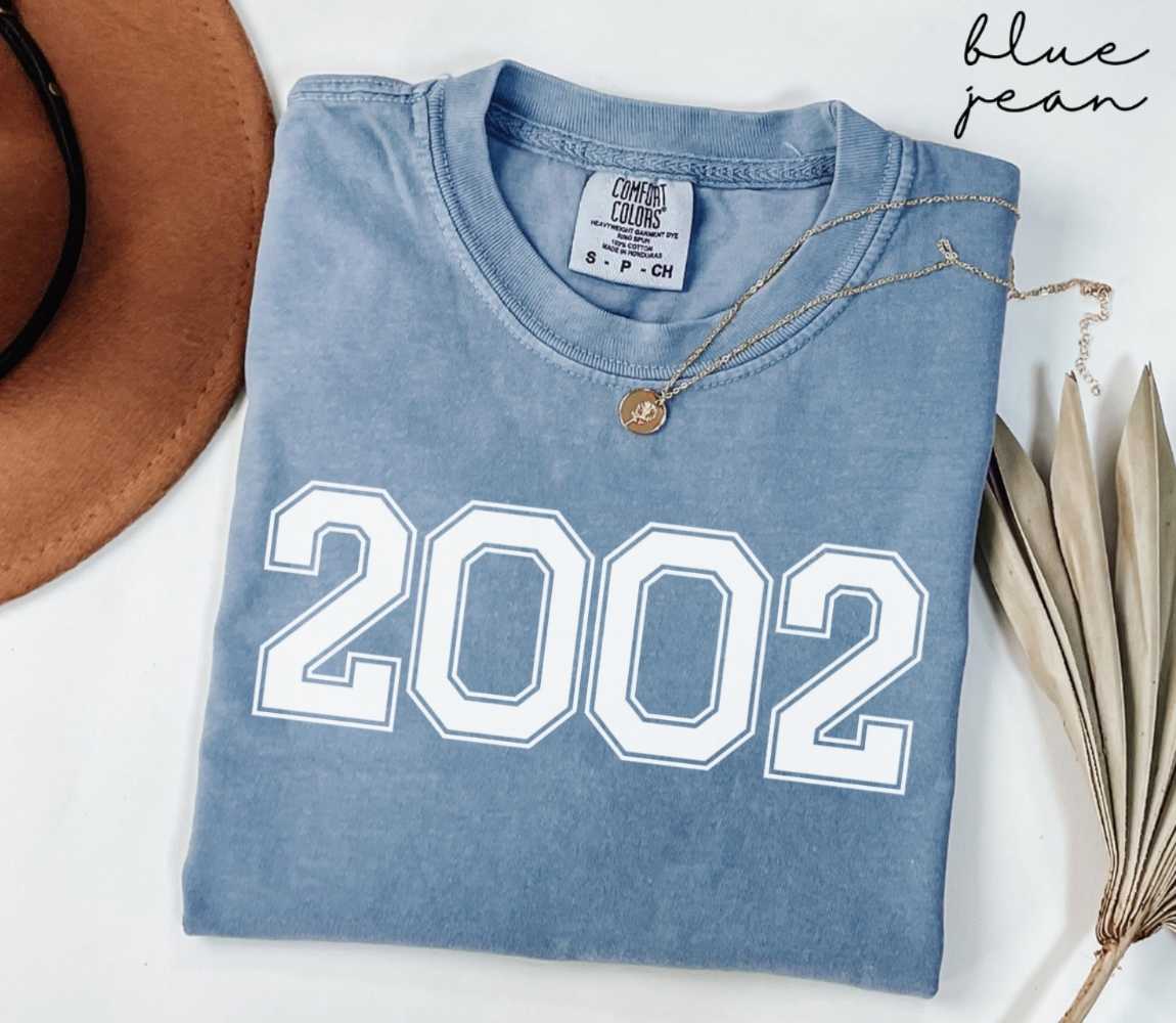 Folded blue jean t-shirt featuring '2002' print on a white surface, accompanied by a hat and dried leaves.
