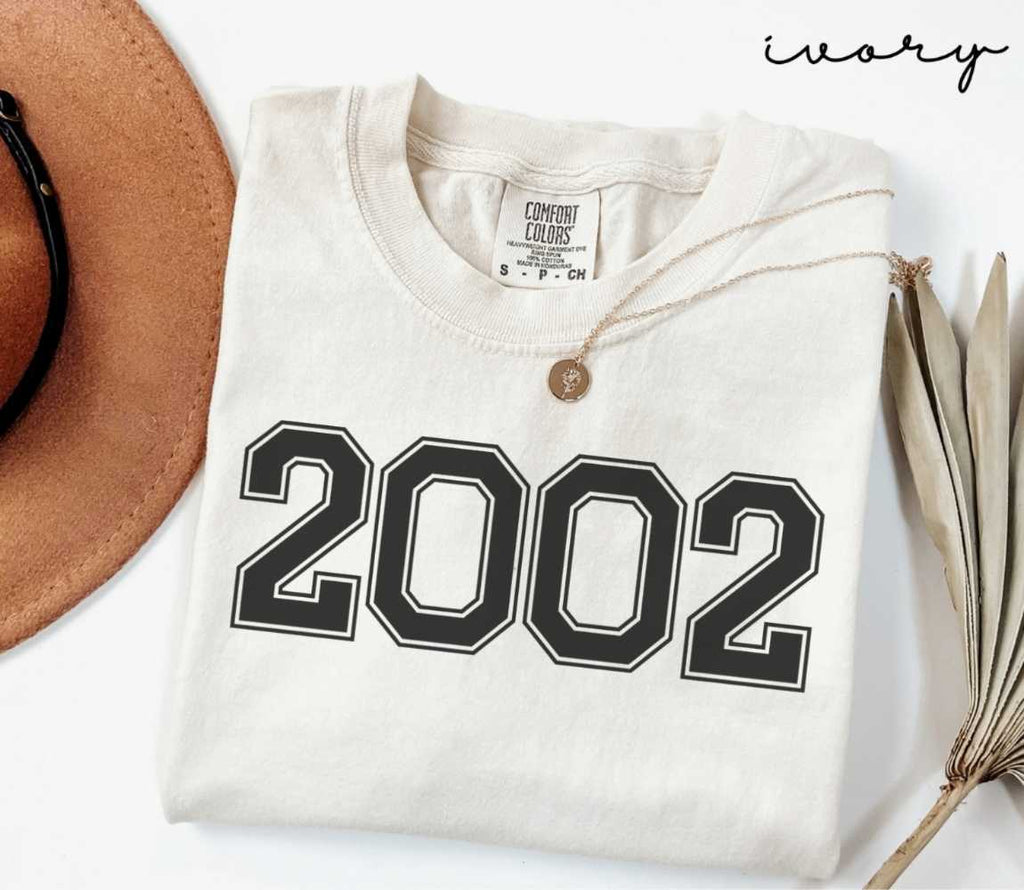 A beige (ivory) t-shirt with '2002' print, placed next to a brown hat and a dried plant on a light background.
