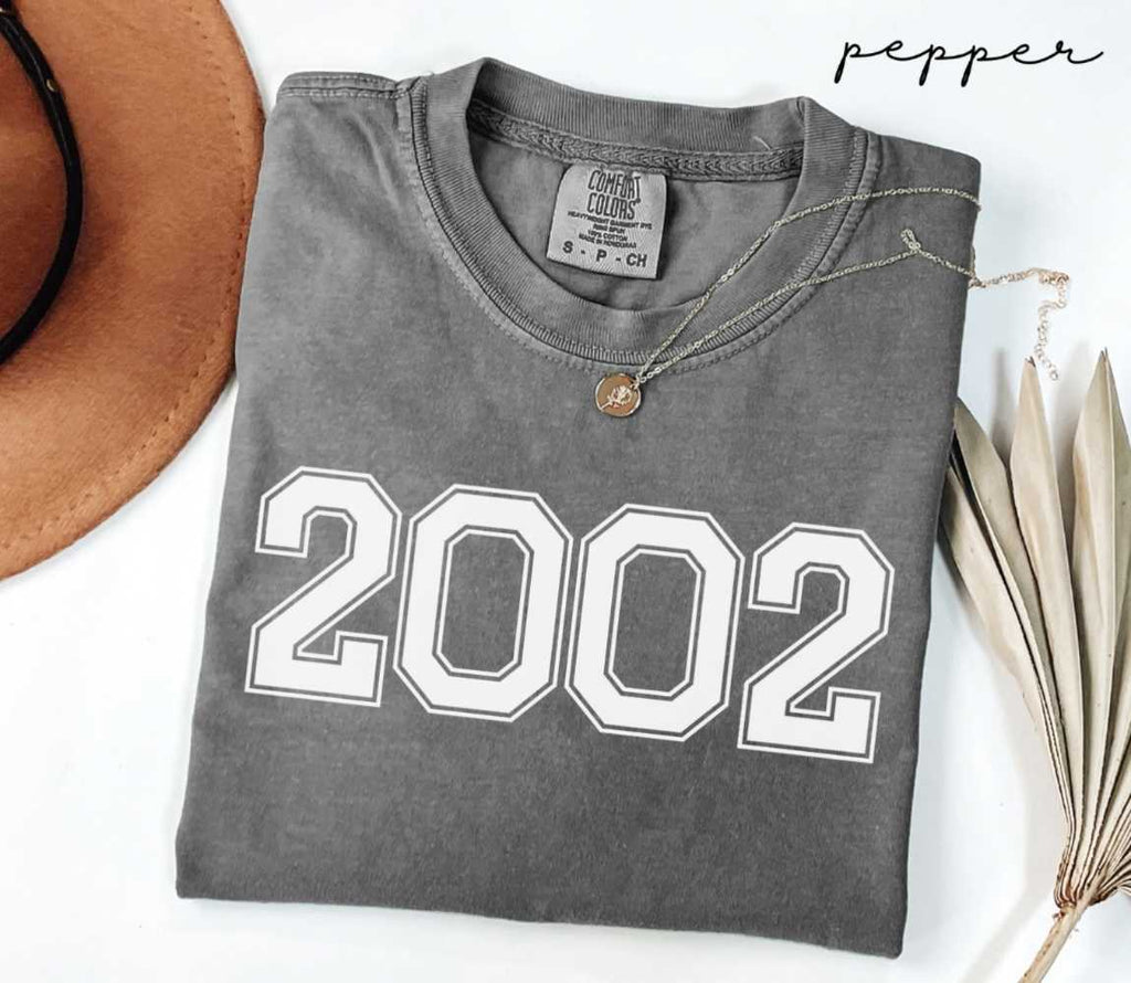 Folded gray (pepper) t-shirt with '2002' print on a white surface with a hat and dried leaves.
