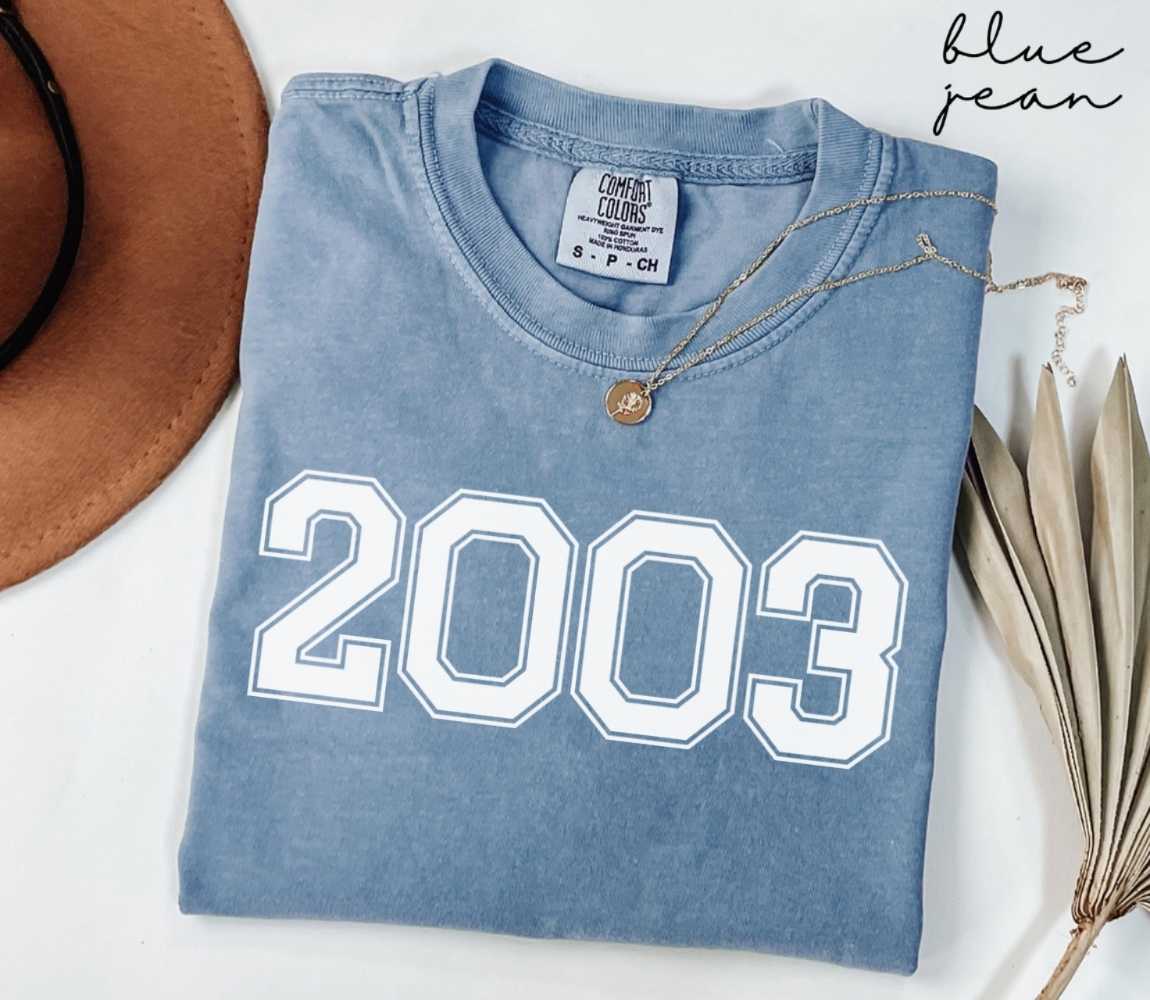 Folded blue jean t-shirt featuring '2003' print on a white surface, accompanied by a hat and dried leaves.
