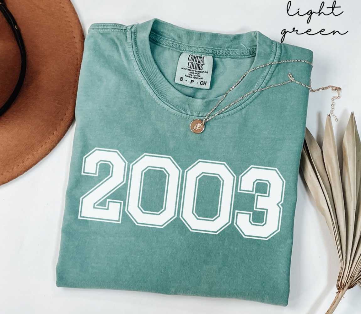 A light green t-shirt with '2003' printed on it, placed on a white surface, accompanied by a hat and dried leaves.
