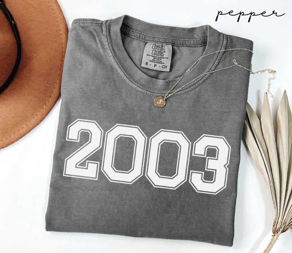 Folded gray (pepper) t-shirt with '2003' print on a white surface with a hat and dried leaves.
