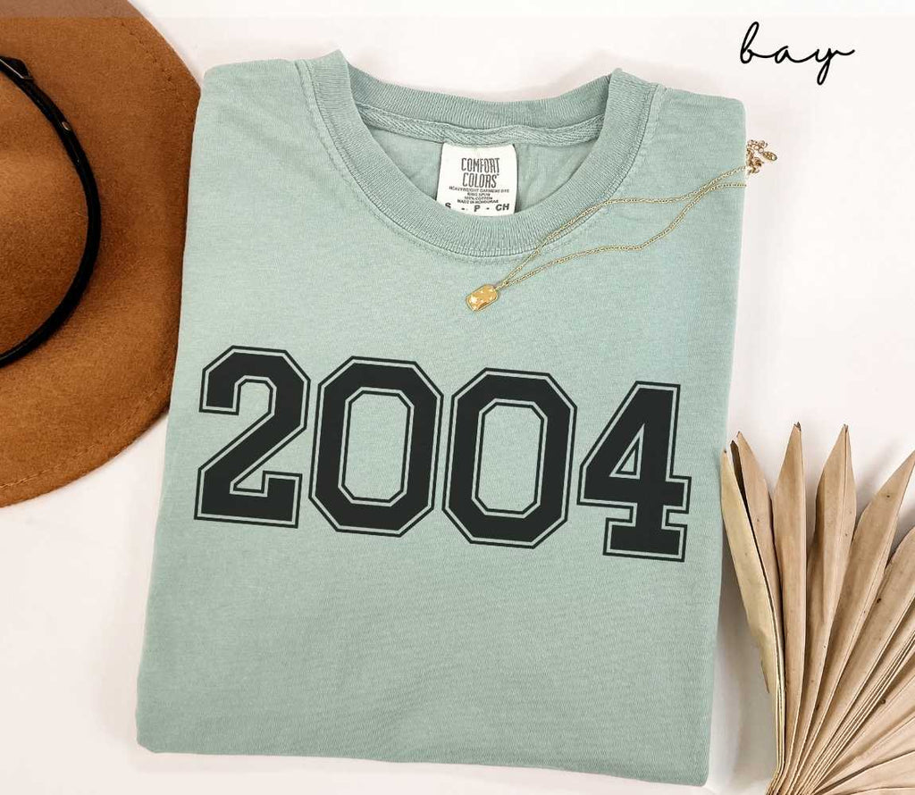A light green (bay) t-shirt with "2004" printed on it, placed on a white surface alongside a hat and dried leaves.
