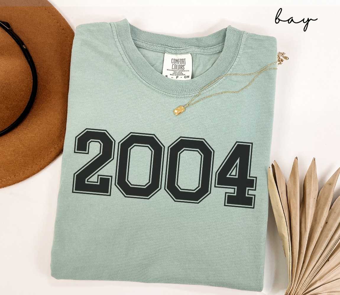A light green (bay) t-shirt with "2004" printed on it, placed on a white surface alongside a hat and dried leaves.
