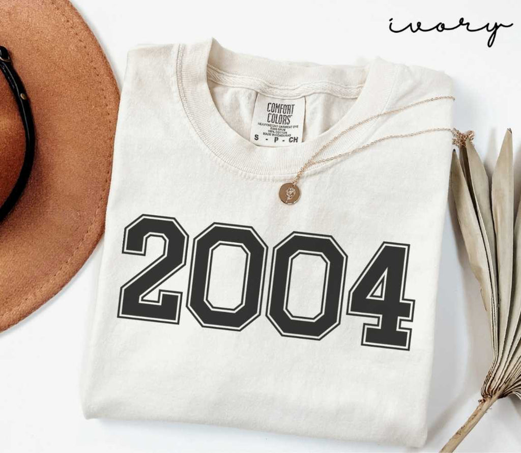 A beige (ivory) t-shirt with '2004' print, placed next to a brown hat and a dried plant on a light background.
