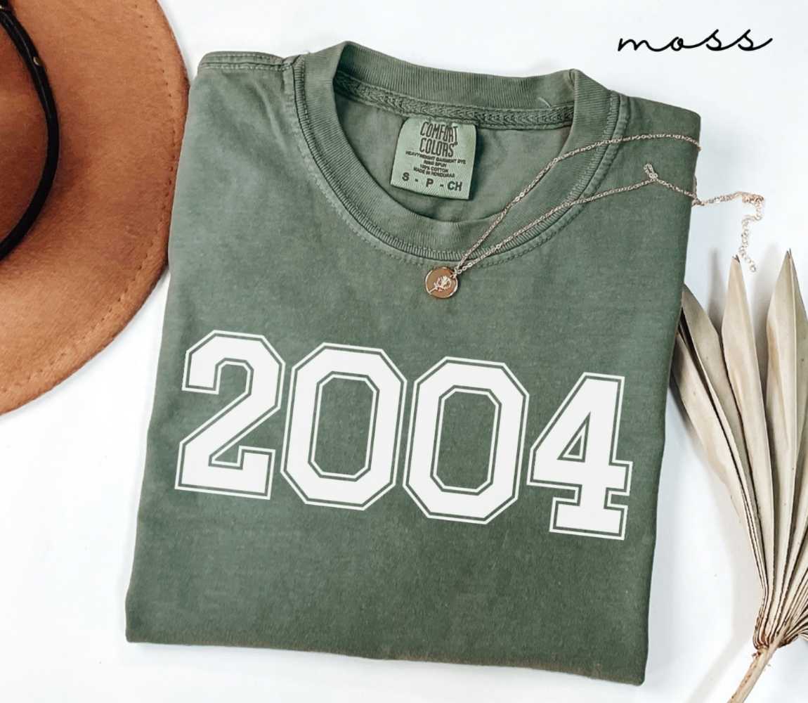Folded green (moss) t-shirt featuring a '2004' print on a white surface alongside a hat and dried leaves.
