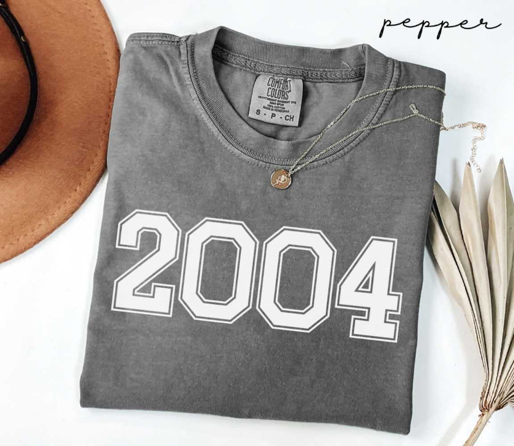 Folded gray (pepper) t-shirt with '2004' print on a white surface with a hat and dried leaves.
