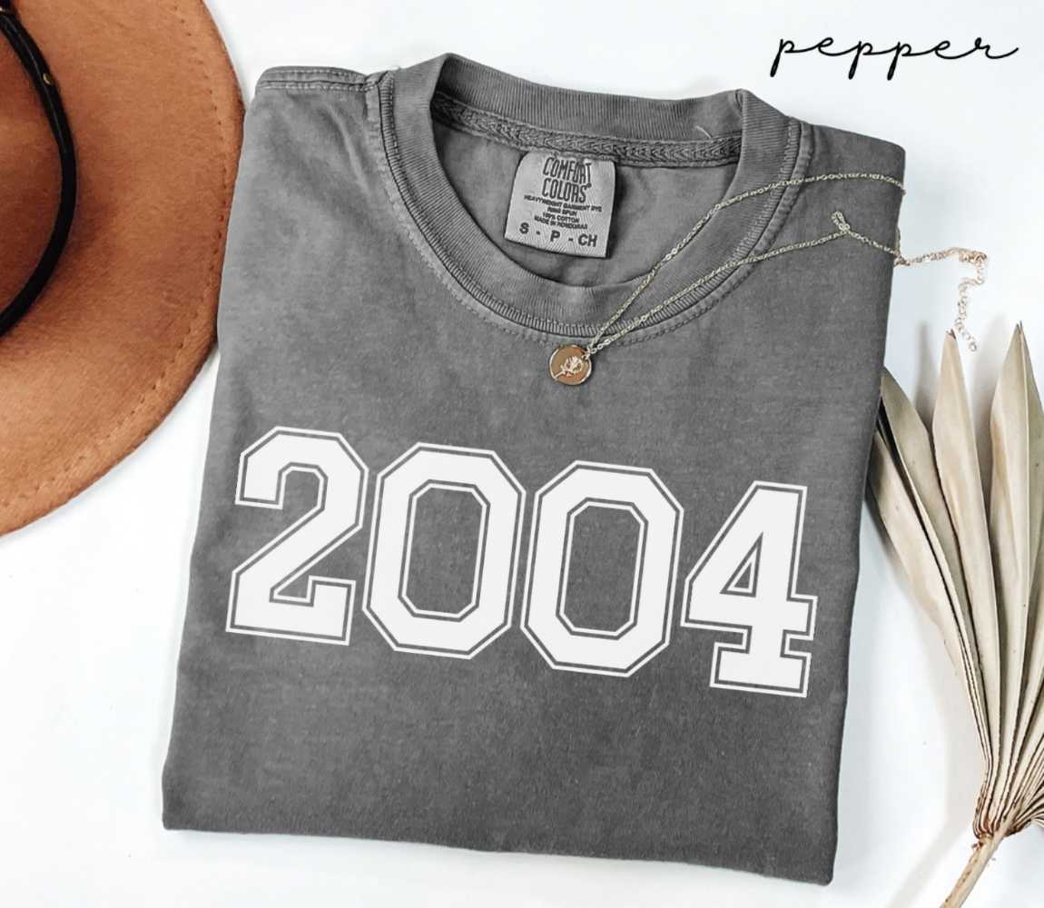 Folded gray (pepper) t-shirt with '2004' print on a white surface with a hat and dried leaves.
