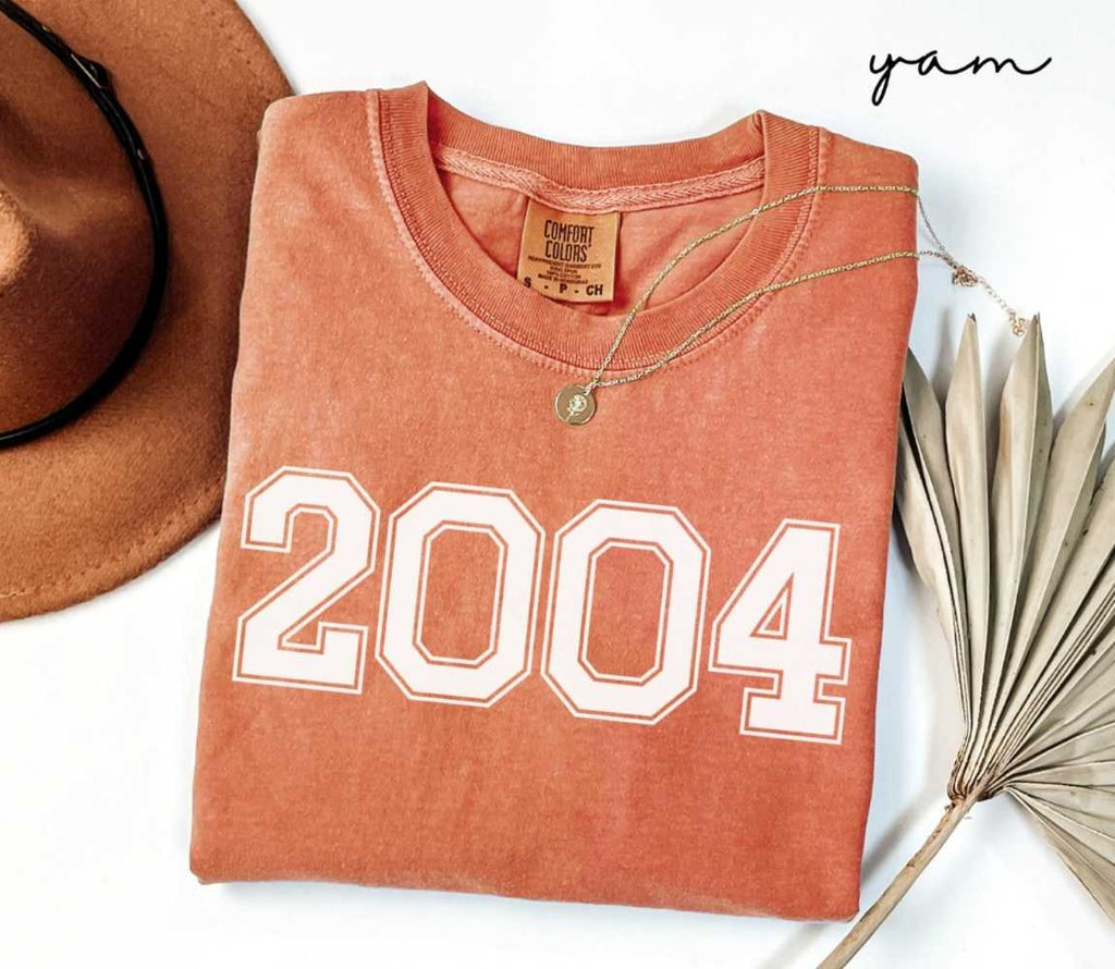 Folded orange (yam) t-shirt with a '2004' print, a brown hat, and a dried plant on a white background.
