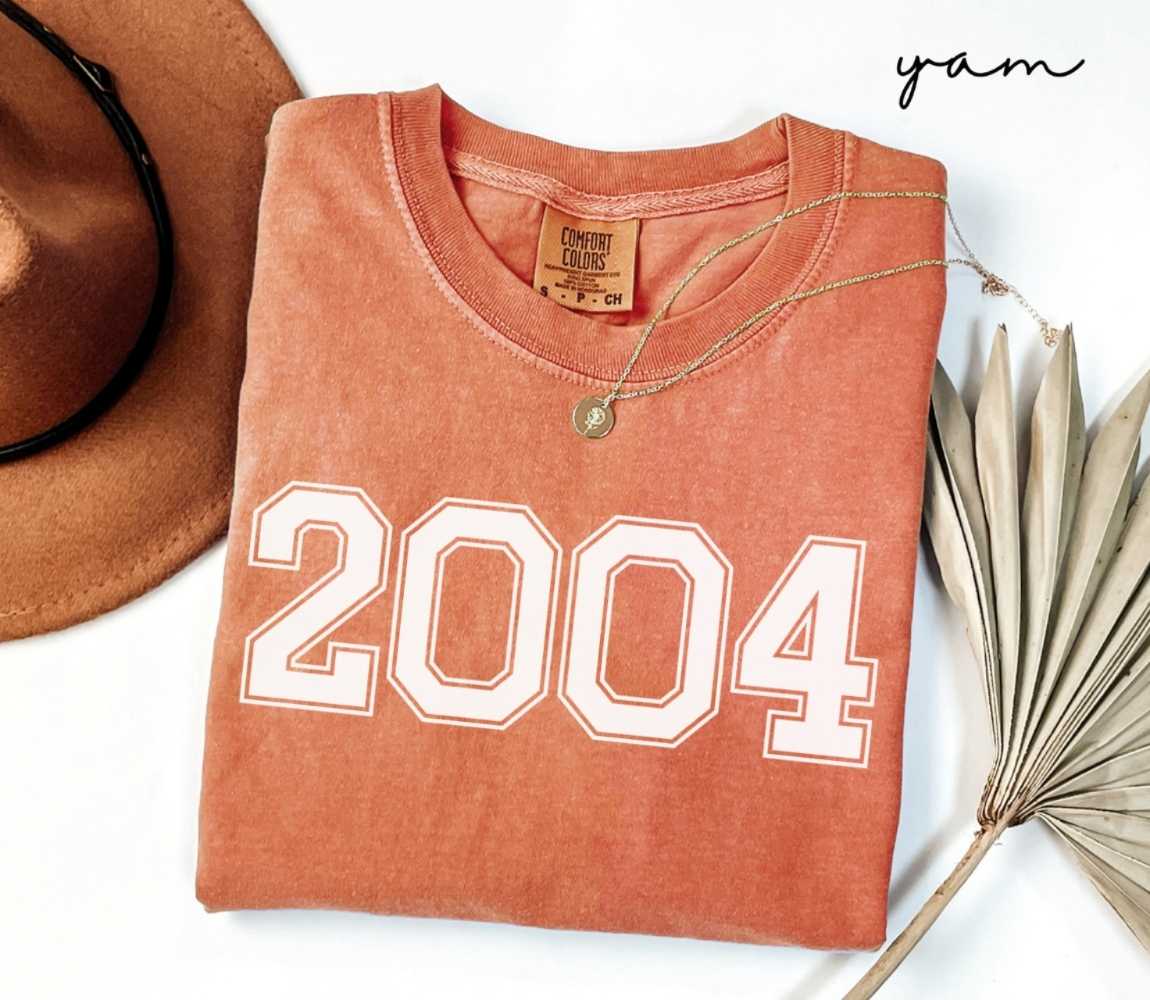 Folded orange (yam) t-shirt with a '2004' print, a brown hat, and a dried plant on a white background.
