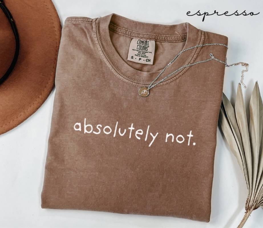 Folded brown t-shirt with the text 'Absolutely not.' next to a hat and dried leaves on a white background.