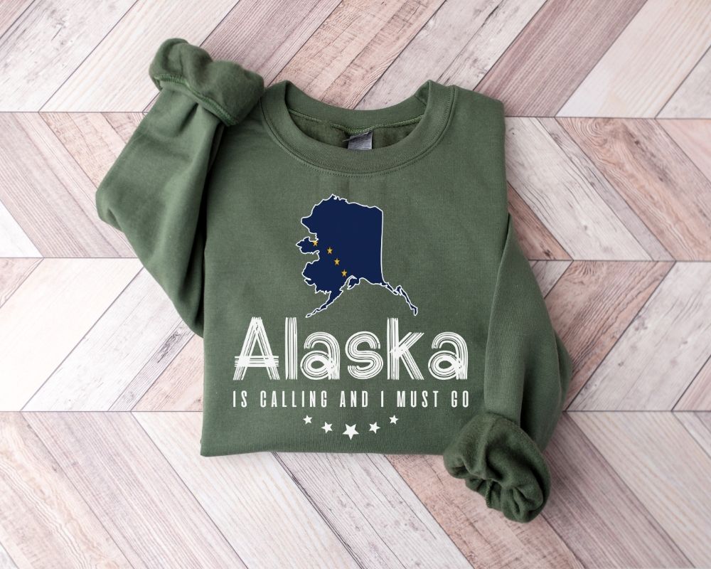 Military green sweatshirt with blue Alaska map and flag outline and text "Alaska is calling I must go" on wooden floor