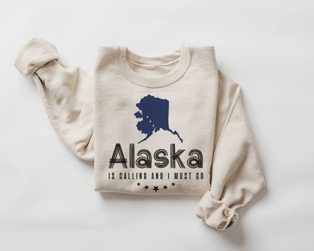 Beige (Sand) sweatshirt with blue Alaska map and flag outline and text "Alaska is calling I must go" on a white background