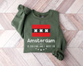 A millitray green sweatshirt featuring the word "Amsterdam" along with a flag design displayed on a wooden floor.