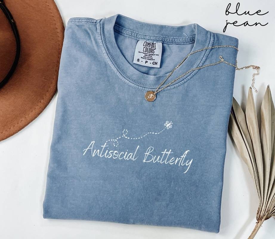 A folded blue jean t-shirt featuring the text "Antisocial Butterfly" is placed next to a hat and dried leaves on a white background.