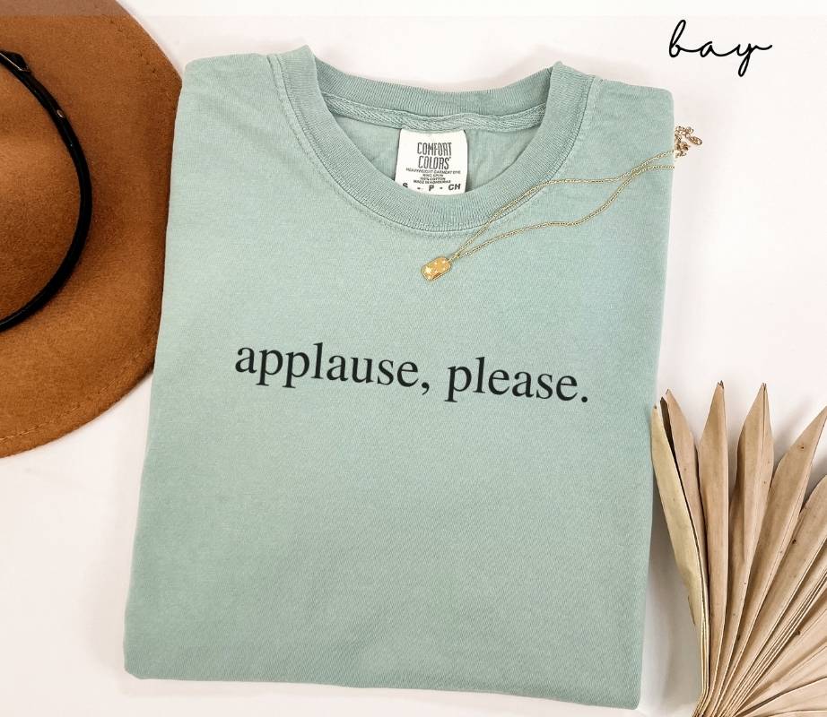 Faded light green (bay) t-shirt featuring the text 'Applause, please,' on a white background, accompanied by a hat and dried leaves.