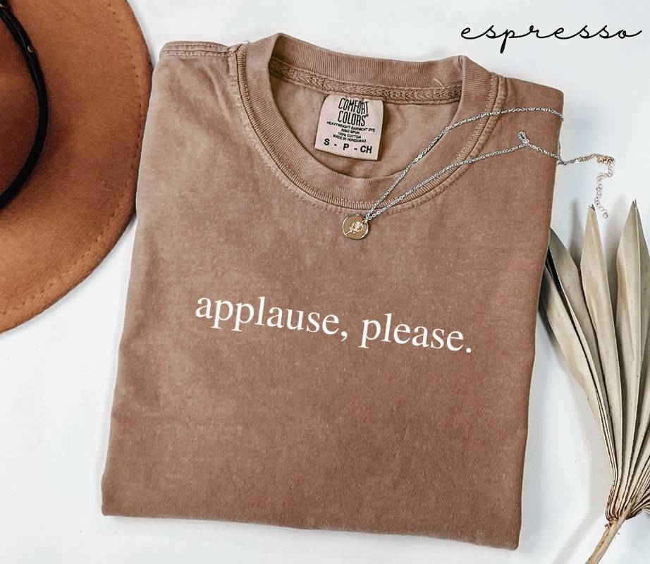 Folded brown (espresso) t-shirt with the text 'Applause, please.' next to a hat and dried leaves on a white background.