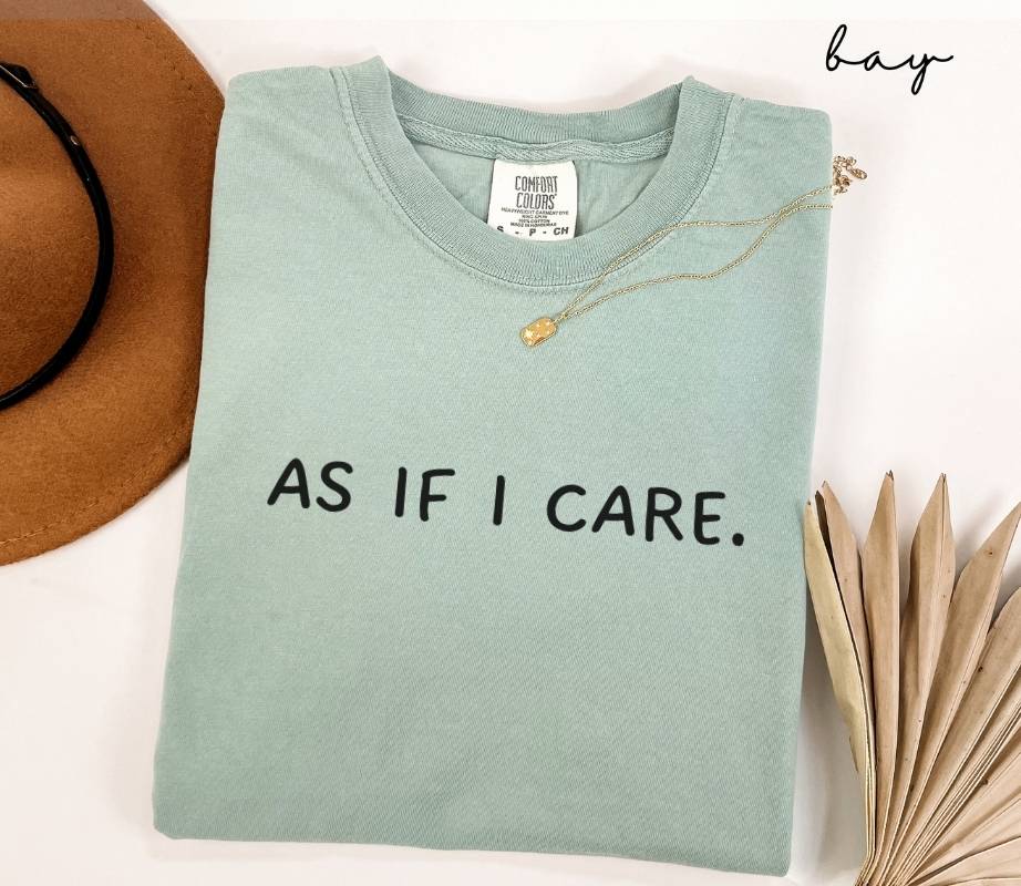 A folded light Green t-shirt with the text 'AS IF I CARE' sits next to a brown hat and dried leaves on a white background.
