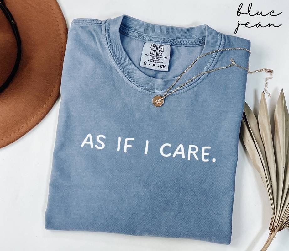 A folded blue jean t-shirt with "AS IF I CARE." text, beside a hat and dried leaves on a white background.