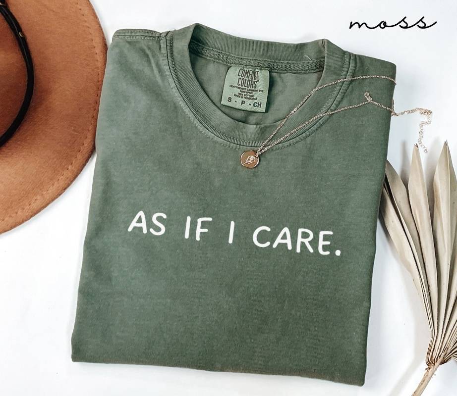 Folded green t-shirt with the text 'AS IF I CARE.' next to a hat and dried leaves on a white background.