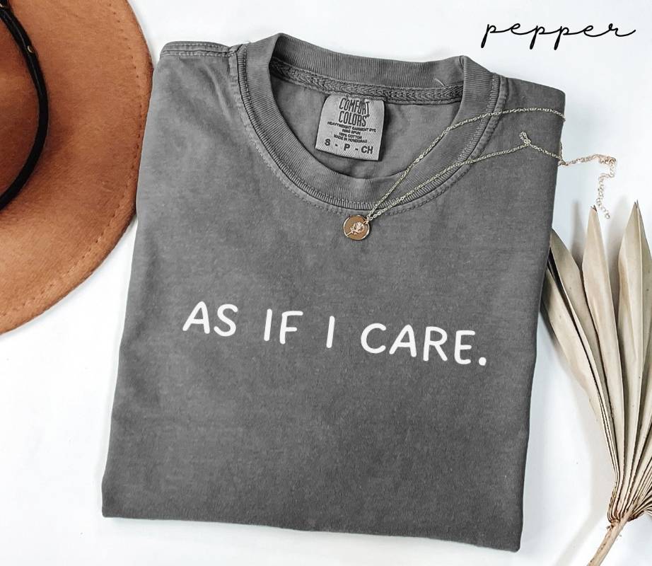 Gray folded t-shirt featuring the text 'AS IF I CARE.' set against a white background, accompanied by a hat and a plant.