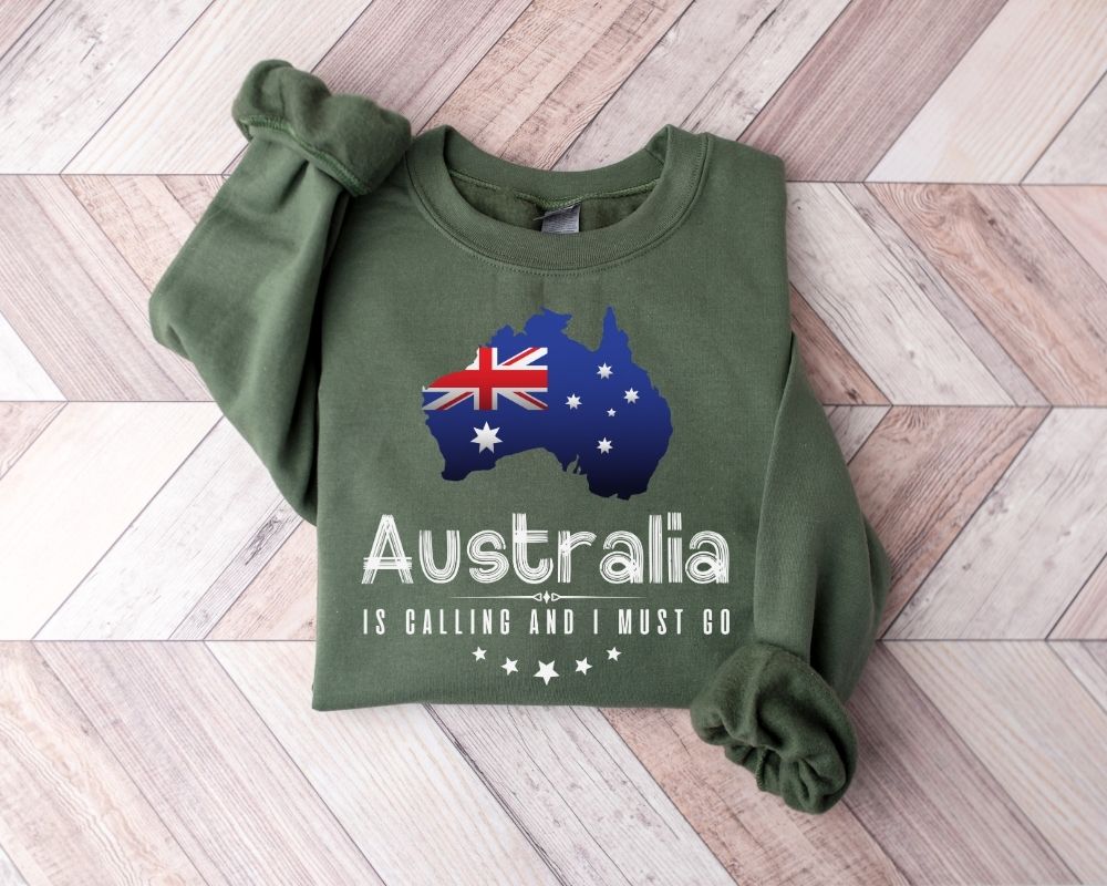 A military green sweatshirt featuring a design of the map and flag of Australia is laid out on a wooden floor.