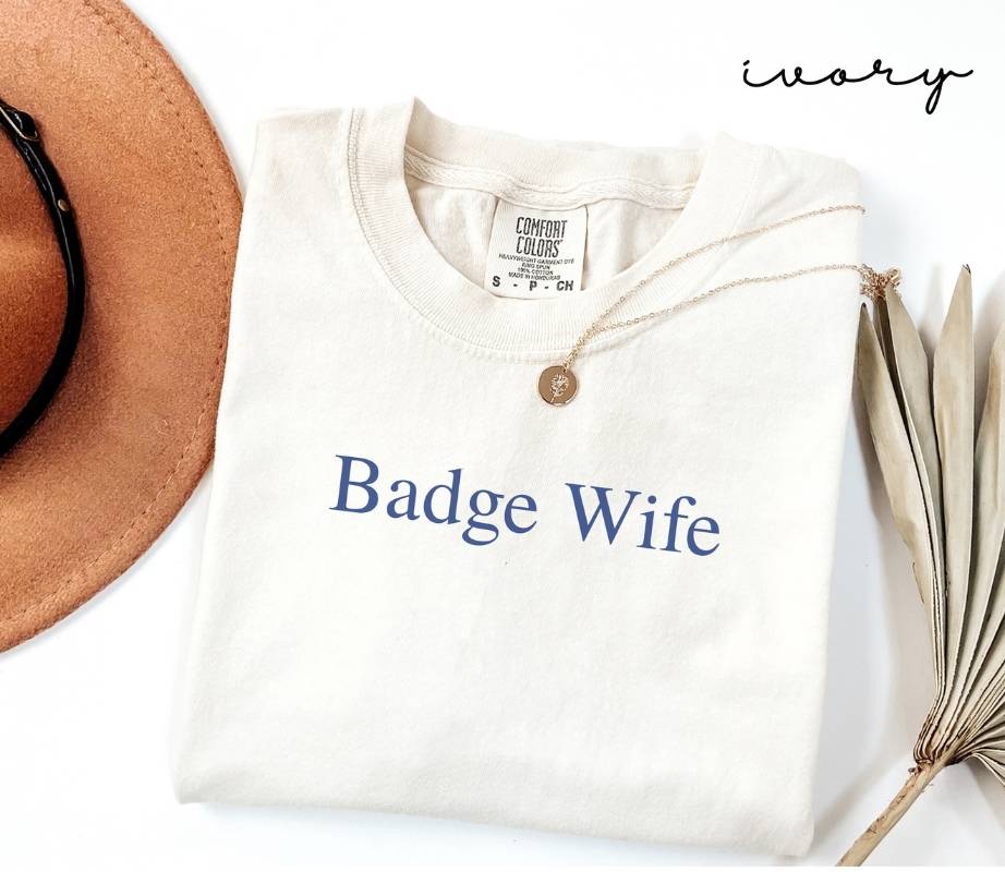 White (ivory) t-shirt with 'Badge Wife' text, brown hat, and dried plant on a white background