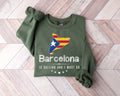 A millitray green sweatshirt featuring a Barcelona design is placed on a wooden floor.