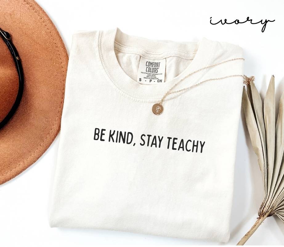 White (ivory) t-shirt with 'BE KIND, STAY TEACHY' text, hat, necklace, and dried plant on a white background