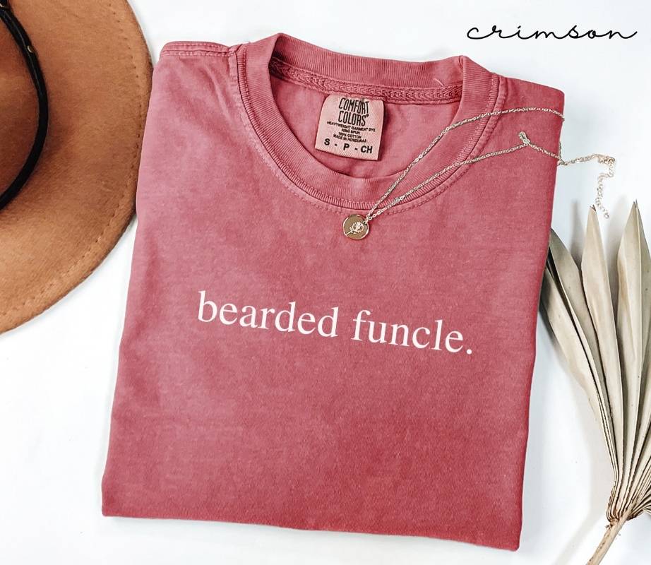 Folded red t-shirt with the text 'bearded funcle,' next to a hat and dried leaves on a white background.