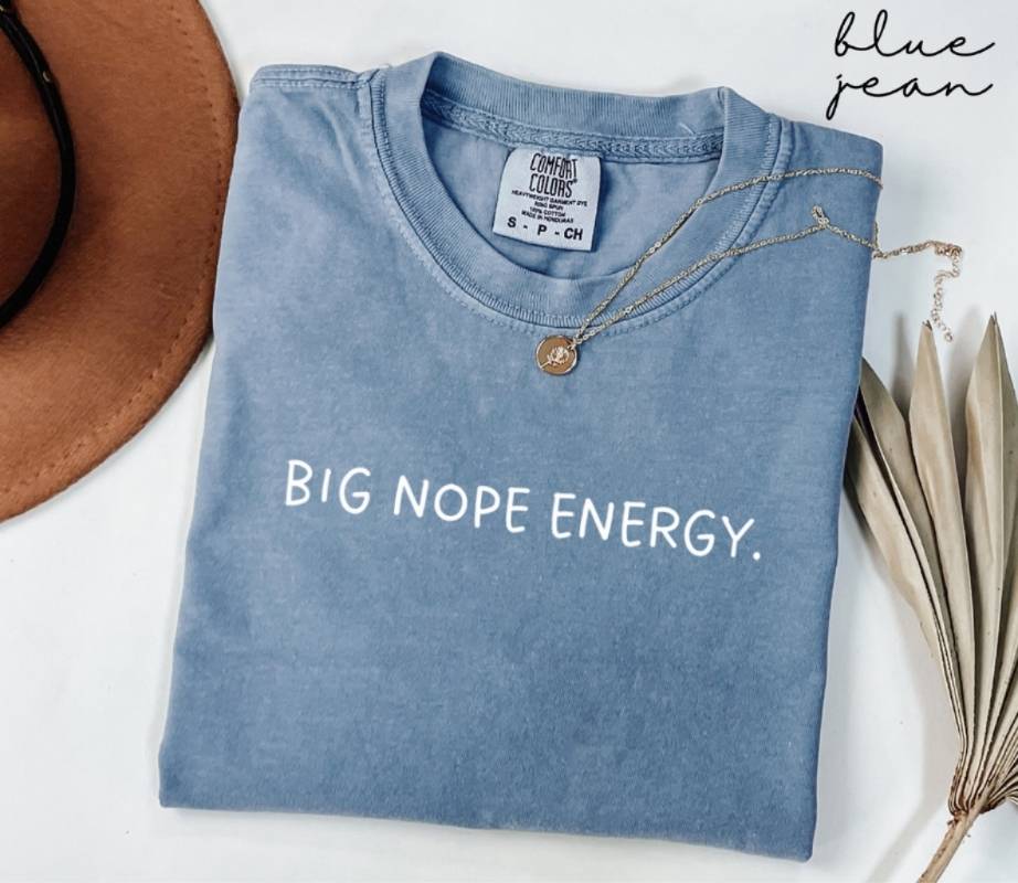 A blue T-shirt with the text 'BIG NOPE ENERGY', beside a hat and dried leaves on a white background.