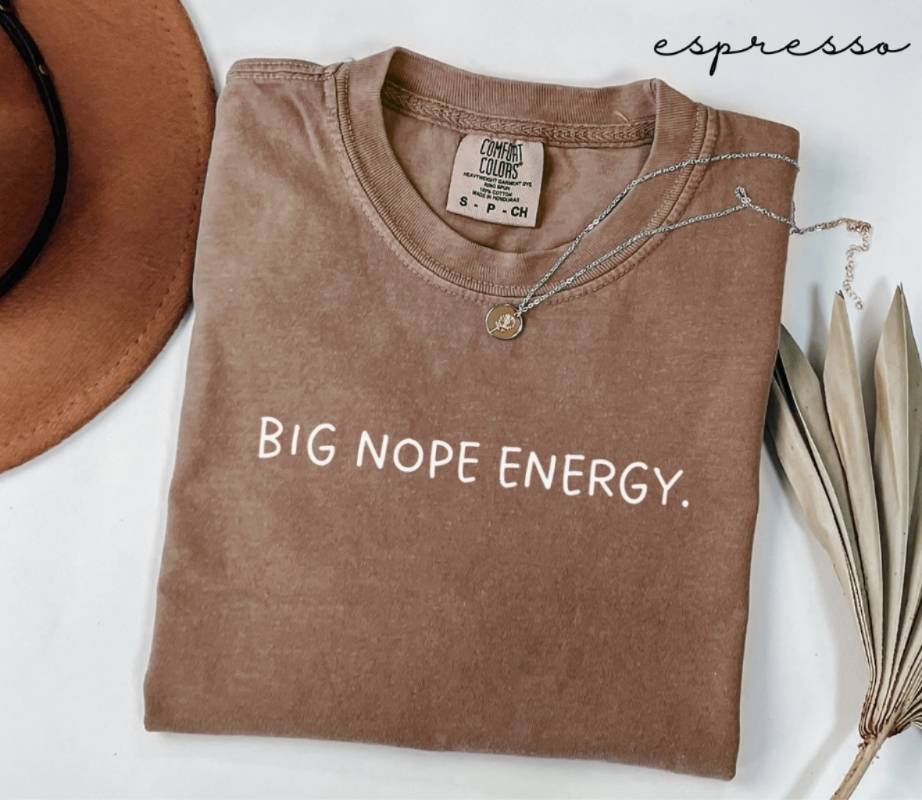 Brown t-shirt with 'BIG NOPE ENERGY.' text, next to a hat and dried leaves on a white background.