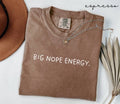 Brown t-shirt with 'BIG NOPE ENERGY.' text, next to a hat and dried leaves on a white background.