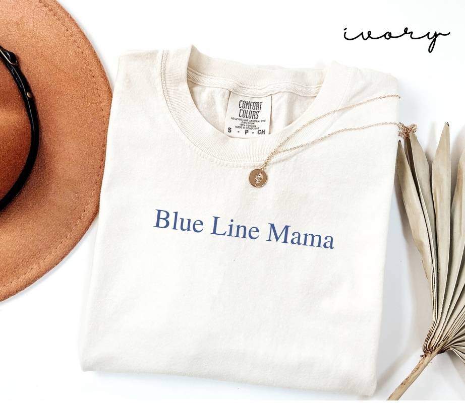 White (ivory) t-shirt with 'Blue Line Mama' text, brown hat, and dried plant on a white background