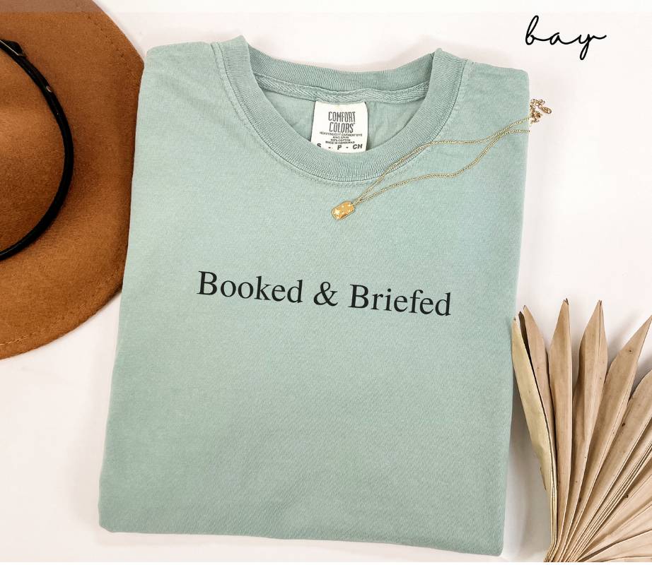 Folded light green (bay) t-shirt with 'Booked & Briefed' text, next to a hat and dried leaves on a white background.