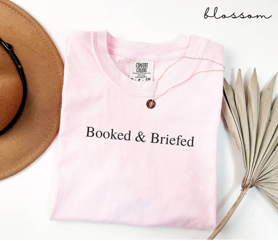 Folded pink (blosoom) t-shirt with 'Booked & Briefed' text, next to a hat and dried plant on a white background.