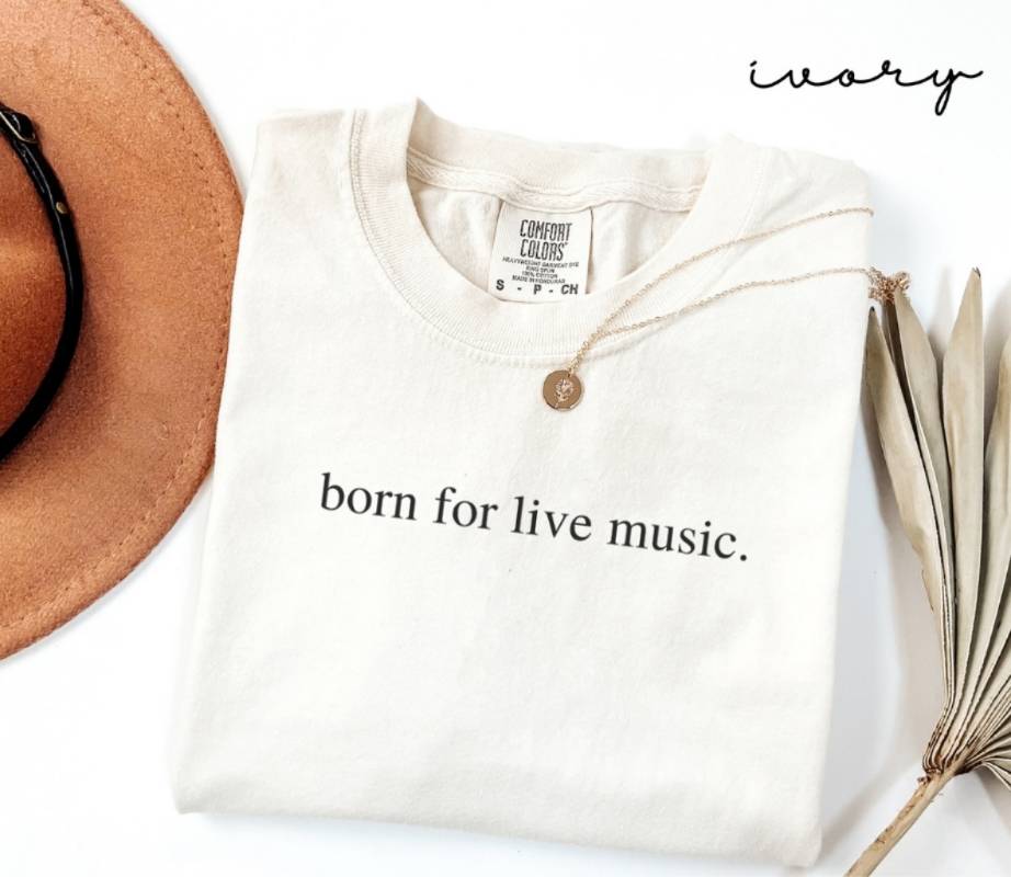 White (ivory) t-shirt with 'born for live music.' text, next to a hat and dried leaves on a white background.