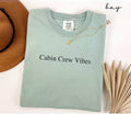 Folded light green (bay) t-shirt with 'Cabin Crew Vibes' text, next to a brown hat and dried leaves on a white background.