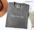 Folded gray (pepper) t-shirt with 'Cabin Crew Vibes' text on a white surface with a hat and dried leaves.