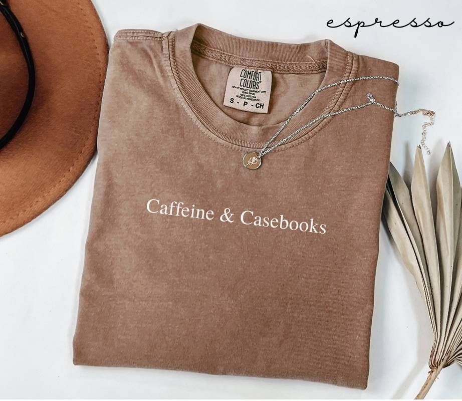Folded brown (espresso) t-shirt with 'Caffeine & Casebooks' text, next to a hat and dried leaves on a white background.