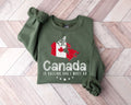 A military green sweatshirt featuring a map of Canada and accompanying text, placed on a wooden floor.