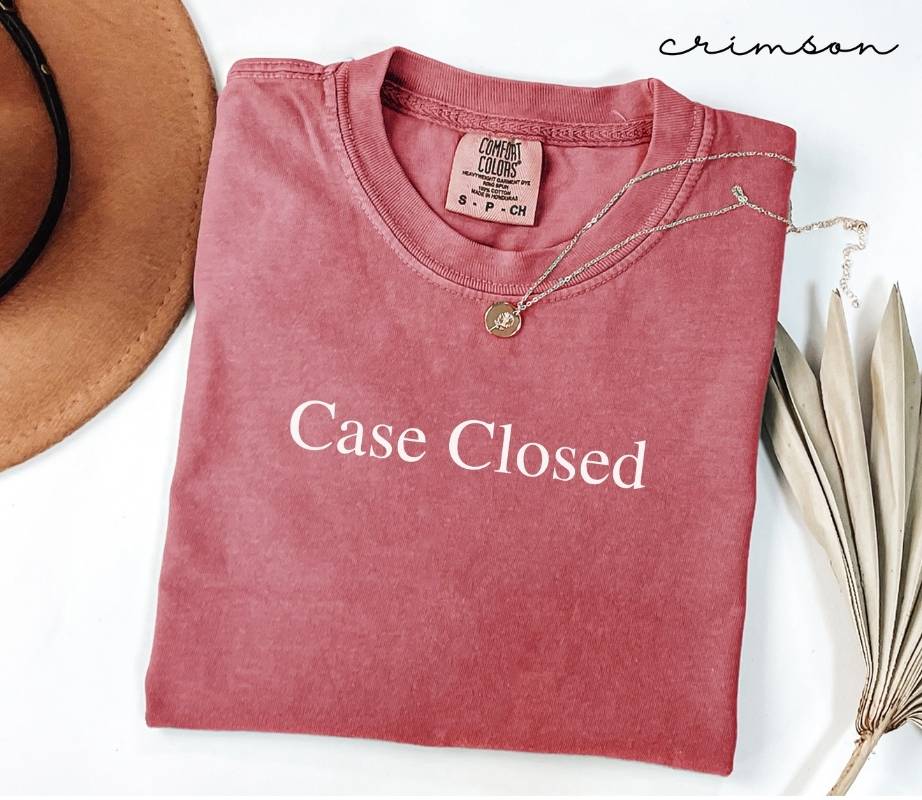 Folded pink (crimson) t-shirt with 'Case Closed' text, necklace, and hat on a white background